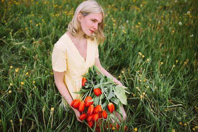 Conteúdo fêmea de vestido em pé com um monte de flores vermelhas de tulipa no prado no verão — Fotografia de Stock