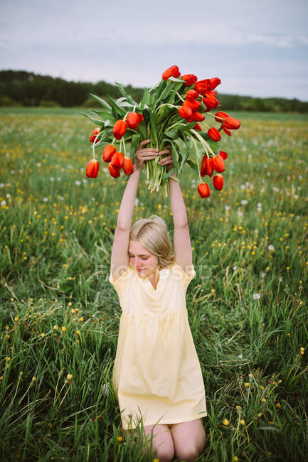 Smiling charming female with bouquet of red tulip flowers sitting in green meadow in summer — Stock Photo