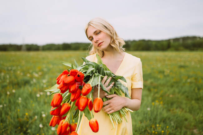 Zufriedene Frau im Kleid steht im Sommer mit einem Strauß roter Tulpenblumen auf der Wiese und schaut weg — Stockfoto
