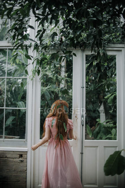Back view of unrecognizable female with long ginger hair reaching hand to door of greenhouse with plants — Stock Photo