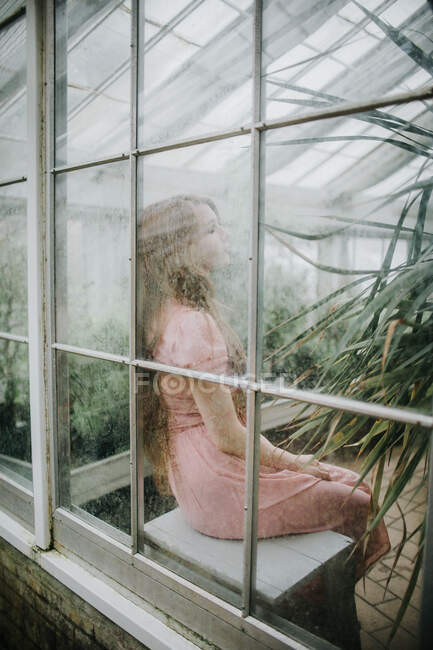 Через стеклянный боковой вид молодой девушки в платье, сидящей в теплице с зелеными растениями — стоковое фото