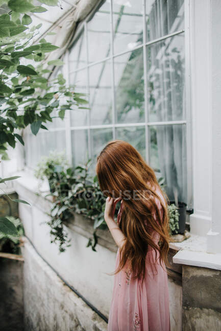 Femme rousse rêveuse méconnaissable en robe debout près des fenêtres en verre de la serre avec des plantes luxuriantes — Photo de stock