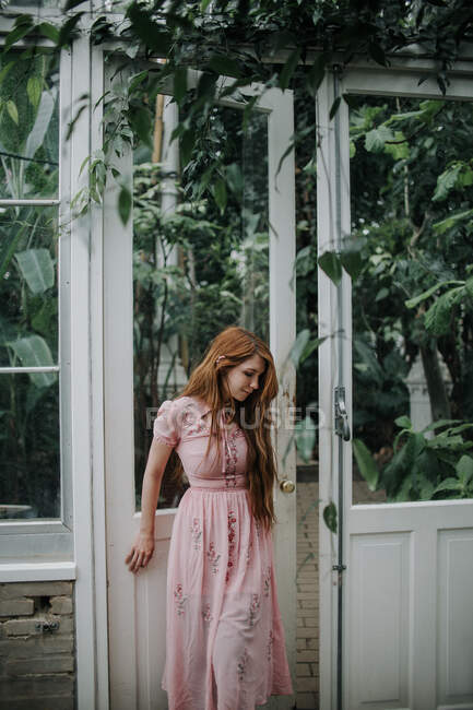 Dreamy redhead female with long hair opening door of greenhouse with plants looking down — Stock Photo