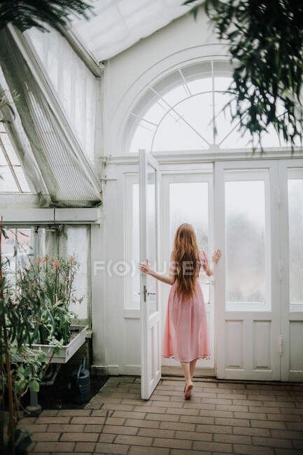 Вид ззаду на невпізнавану жінку з довгим імбирним волоссям, що відкриває двері теплиці з рослинами — стокове фото