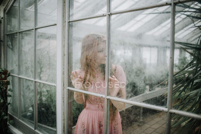 Através de vidro de serena jovem fêmea de vestido em pé na estufa com plantas verdes — Fotografia de Stock
