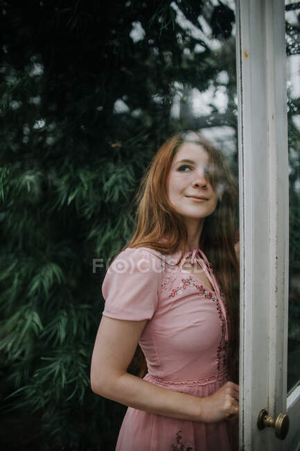 Verträumte positive rothaarige Frau im Kleid steht in der Nähe der Glastür eines Gewächshauses mit üppigen Pflanzen — Stockfoto