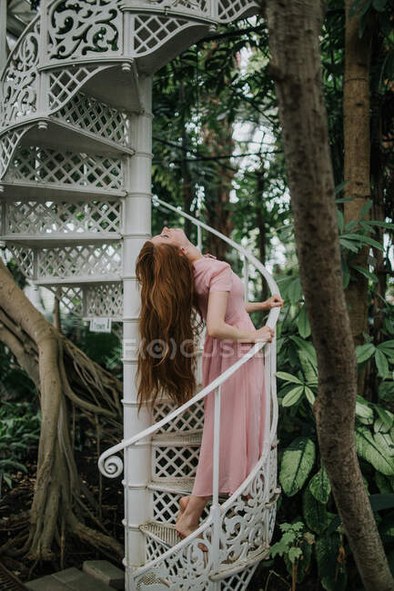 Side view full body of female with long hair standing near railing of stairway in glasshouse — Stock Photo