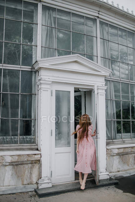 Full body of anonymous female in stylish outfit standing on street in doorway of hothouse with glass walls and growing green plants — Stock Photo