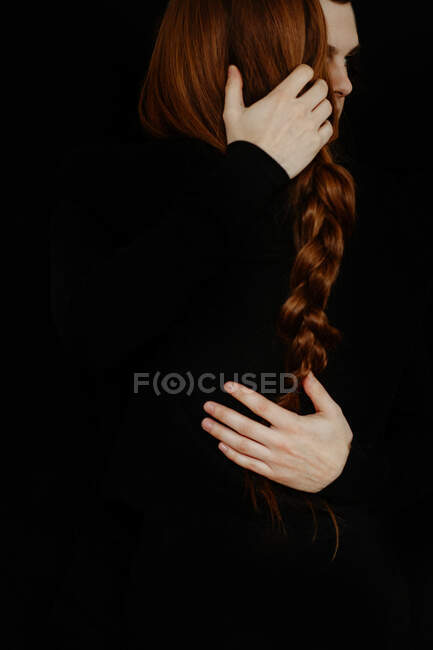 Back view of unrecognizable crop tender boyfriend tenderly embracing redhead girlfriend while standing in dark studio on black background — Stock Photo