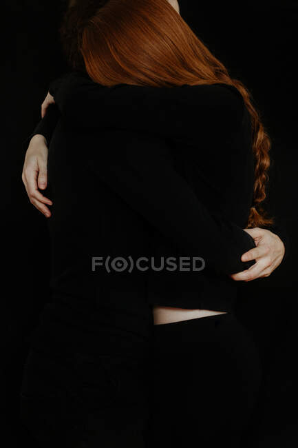 Unrecognizable side view of cropped tender boyfriend tenderly embracing redhead girlfriend while standing in dark studio on black background — Stock Photo
