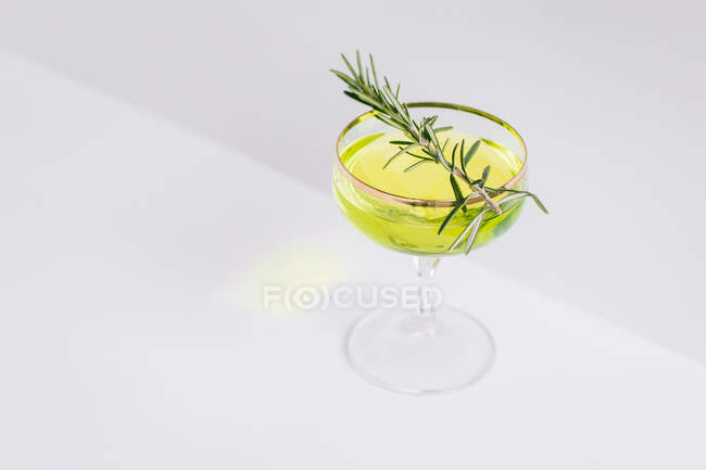 Alto angolo di rinfrescante cocktail alcolico giallo guarnito con rametto di rosmarino fresco posto sul tavolo bianco in studio — Foto stock