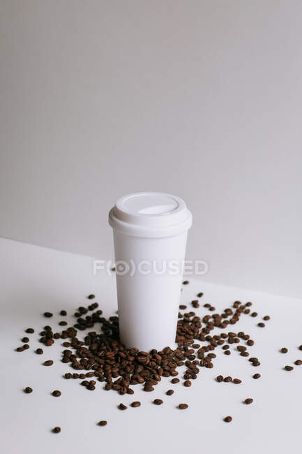 Alto ângulo de taça de papel takeaway colocado na mesa branca com grãos de café espalhados em estúdio — Fotografia de Stock