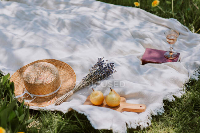 Chapéu de palha feminino e flores colocadas perto de peras frescas em manta de piquenique com livro e copo de vinho no prado de verão verde — Fotografia de Stock