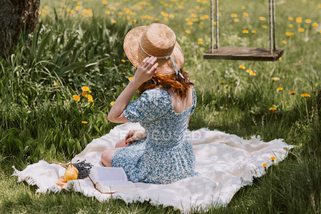 Вид збоку на невпізнавану жінку в сукні та солом'яному капелюсі, що сидить на пікніку на зеленому лузі біля гойдалок у літній сільській місцевості — стокове фото