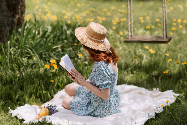 Seitenansicht einer nicht wiedererkennbaren Frau in Kleid und Strohhut, die Roman liest, während sie auf einer Picknickdecke auf einer grünen Wiese in der Nähe von Schaukeln in der sommerlichen Landschaft sitzt — Stockfoto