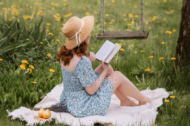 Vue latérale de la femelle méconnaissable en robe et chapeau de paille lecture roman tout en étant assis sur la couverture de pique-nique sur prairie verte près des balançoires dans la campagne estivale — Photo de stock