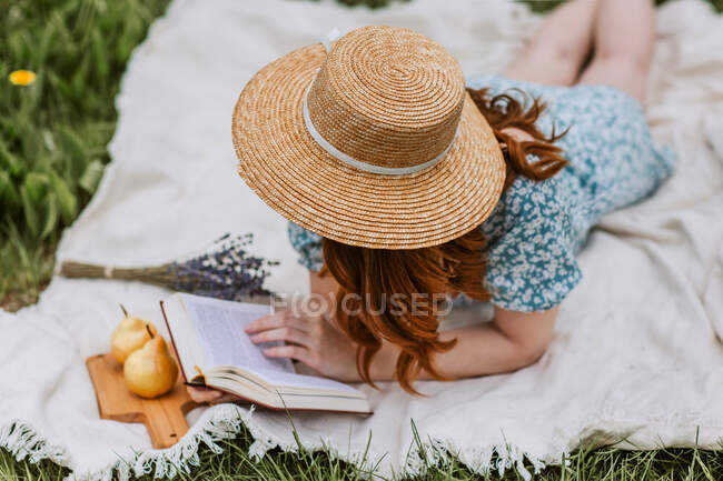 Alto ángulo de hembra irreconocible en vestido con sombrero de paja en la cara acostado con libro abierto en manta de picnic mientras se enfría solo y disfruta del fin de semana de verano en el campo - foto de stock