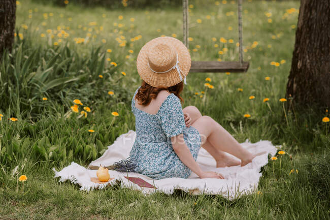 Side view of unrecognizable female in dress and straw hat sitting on picnic blanket on green meadow near swings in summer countryside — Stock Photo