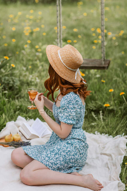 Seitenansicht der nicht wiederzuerkennenden jungen barfüßigen Frau in Sonnenhut und Strohhut entspannt mit einem Glas Getränk auf einer Picknickdecke mit offenem Buch, während sie den Sommertag allein in der Natur genießt — Stockfoto