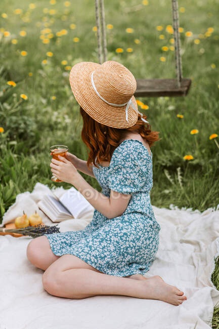 Side view of unrecognizable young barefoot female in sundress and straw hat relaxing with glass of drink on picnic blanket with open book while enjoying summer day alone in countryside — Stock Photo