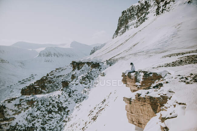 Неузнаваемый космонавт в белом скафандре зимой сидит на краю скалистой горы и любуется удивительным пейзажем Шпицбергена — стоковое фото