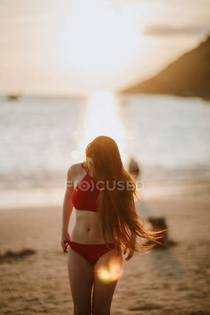 Schlanke Frau mit langen Haaren in rotem Badeanzug steht am Sandstrand vor ruhigem Meer vor verschwommenem Hintergrund in tropischem Land — Stockfoto