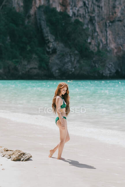 Повний вигляд жінки в купальнику стоїть на піщаному узбережжі біля блакитного моря проти гори в Таїланді. — стокове фото
