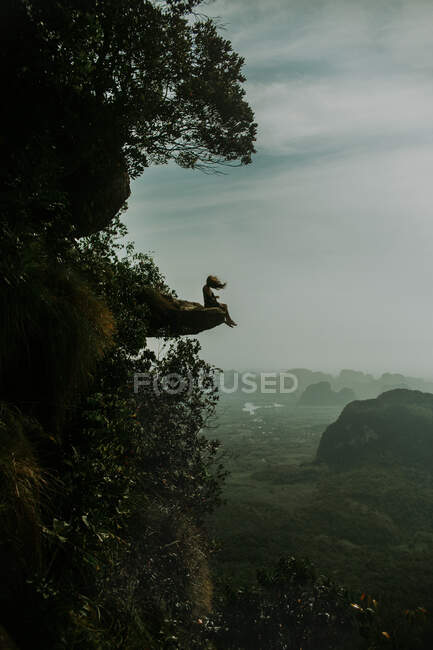 Side view of distant female traveler with flying hair sitting on edge of rock above woodland with green trees in Thailand — Stock Photo