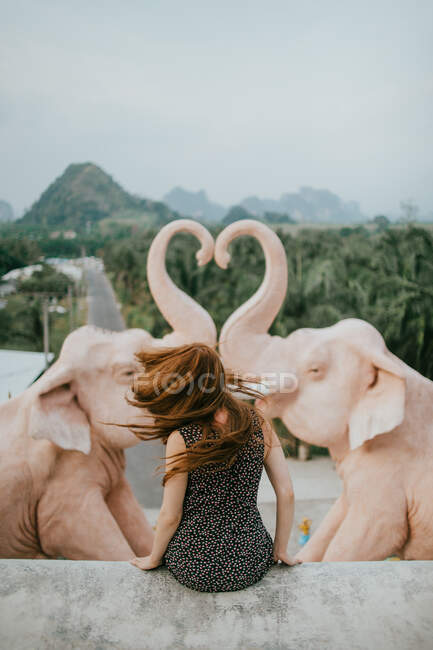 Back view of anonymous female traveler sitting near statue of elephants against lush green trees and mountains in tropical country — Stock Photo