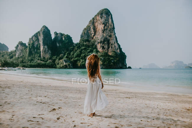 Back view full body of faceless female in white dress standing on sandy beach near azure water against rocky cliffs in Thailand — Stock Photo
