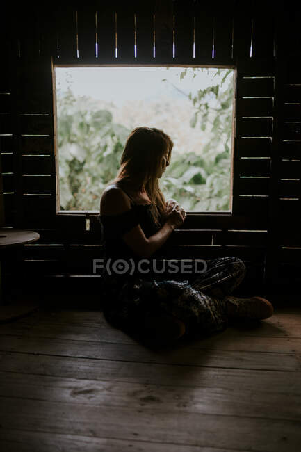 Full body side view of tranquil female sitting in wooden cabin near window in tropical country with lush green plants — Stock Photo