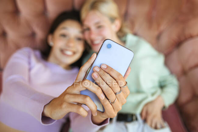 Сверху веселые юные сестры, лежащие на диване и стреляющие в себя по мобильному телефону — стоковое фото