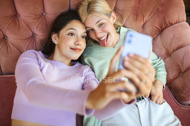 Desde arriba de las jóvenes hermanas alegres tumbadas en el sofá y tomando auto disparo en el teléfono móvil - foto de stock