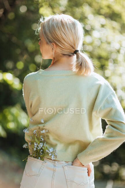 Vue arrière de l'adolescente avec des fleurs sauvages dans la poche du jean debout dans le parc d'été — Photo de stock