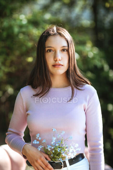 Menina adolescente serena com flores silvestres em jeans em pé no jardim de verão e olhando para a câmera — Fotografia de Stock