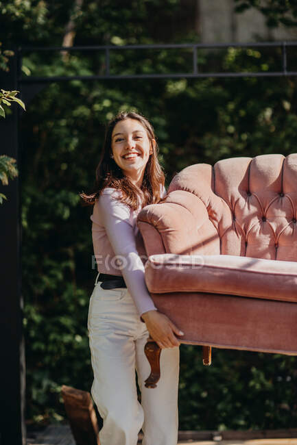 Cheerful teen girl carrying soft couch on terrace in backyard on sunny day and looking at camera — Stock Photo