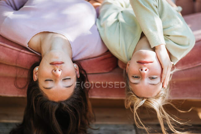 Delicadas irmãs adolescentes deitadas no sofá de cabeça para baixo e desfrutando de dia ensolarado juntos — Fotografia de Stock