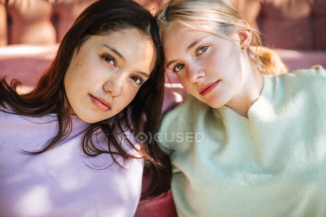 Ніжні сестри-підлітки, сидячи біля зручного дивана в саду в сонячний день і дивлячись на камеру — стокове фото