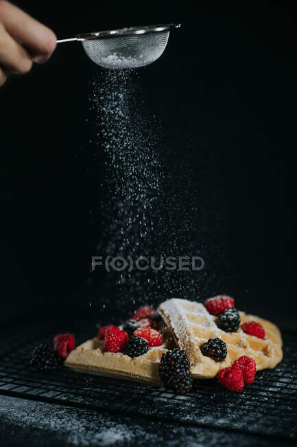 Crop chef anónimo espolvorear azúcar en polvo sobre gofres dulces servidos con bayas frescas sobre fondo negro - foto de stock