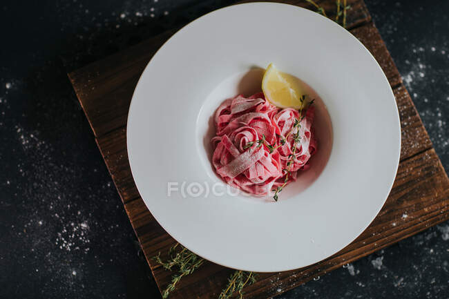 Top view of appetizing vegan pink beetroot pasta with lemon slice and thyme sprig served on wooden board on black table — Stock Photo
