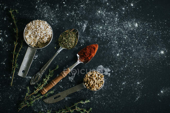 Vista superior de cucharas con pimentón y hierbas secas colocadas sobre mesa negra con semillas de avena y girasol - foto de stock