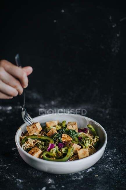 Урожай неузнаваемый человек ест аппетитный вегетарианский салат с жареным тофу и овощами подается на черном фоне — стоковое фото