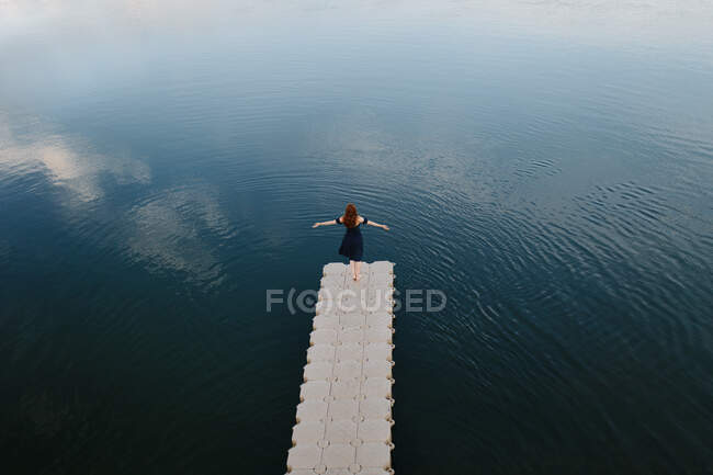 High angle back view of female standing with outstretched arms on quay near lake and enjoying freedom — Stock Photo