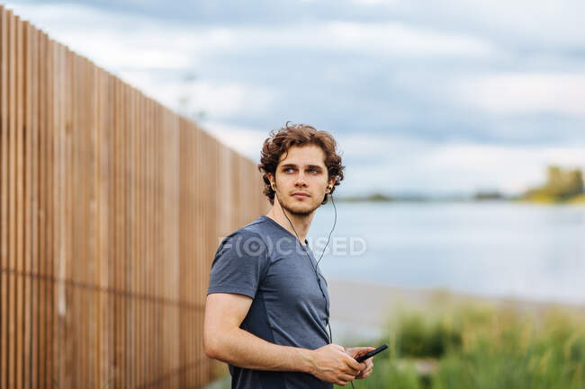 Side view of male athlete listening to music in earphones while standing near lake during training — Stock Photo