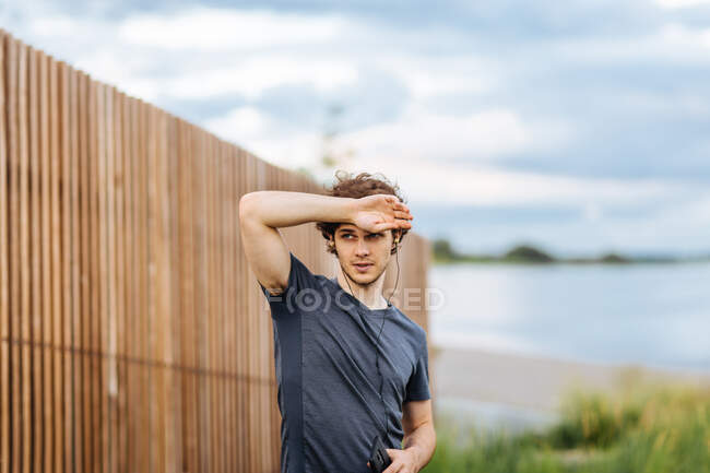 Side view of male athlete listening to music in earphones while standing near lake during training — Stock Photo