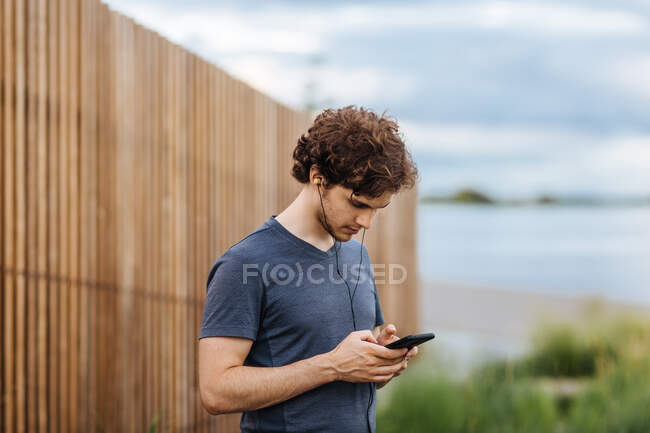 Вид збоку чоловічий спортсмен слухає музику в навушниках, стоячи біля озера під час тренувань — стокове фото