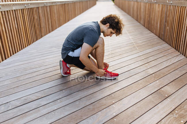 Vista lateral do corredor masculino apto amarrando atacadores em tênis no calçadão de madeira — Fotografia de Stock