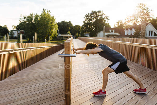 Seitenansicht eines männlichen Athleten, der sich beim Aufwärmen beim Training auf der Holzpromenade im Sommer die Beine streckt — Stockfoto