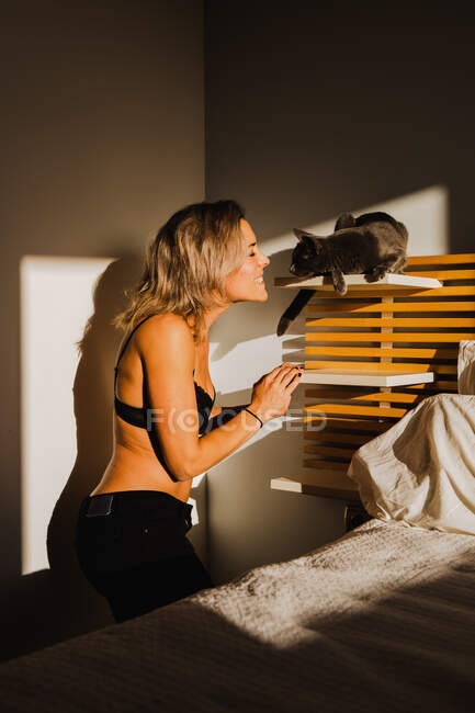 Без сорочки жінка приборкає милий кіт в полиці спальні біля ліжка зі світлом, що потрапляє в кімнату — стокове фото