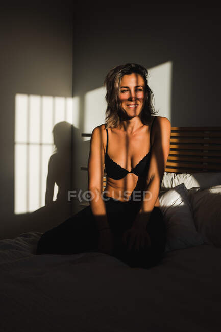 Стрілянина чуттєвої красивої жінки, яка посміхається між світлом і тінями в нижній білизні на ліжку, дивлячись геть — стокове фото